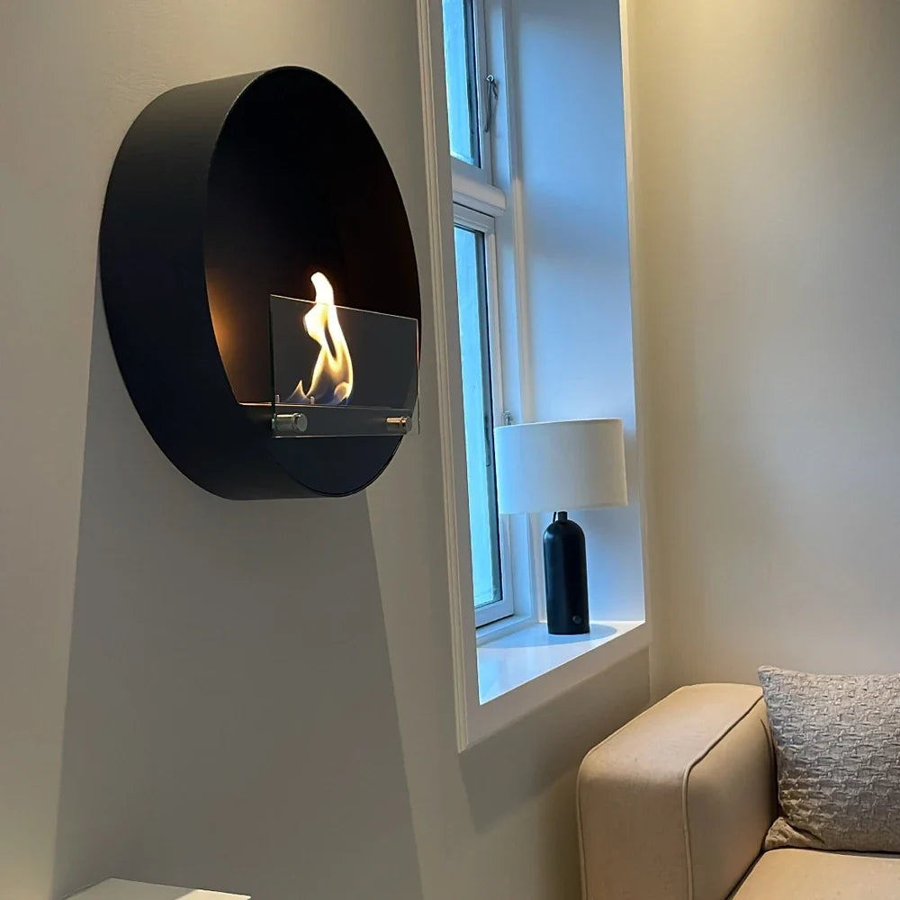 Delaware S black round bio fireplace mounted beside a window nook with a lamp and armchair, lit with a realistic flame